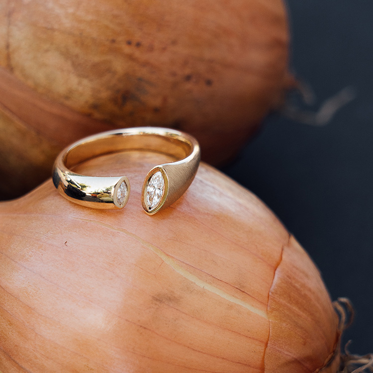 Goldener Ring mit besonderer Form mit Spalt, in die Kanten wurde jeweils ein Brillant im Navette Schliff gefasst. Der Ring liegt auf einer Zwiebel.