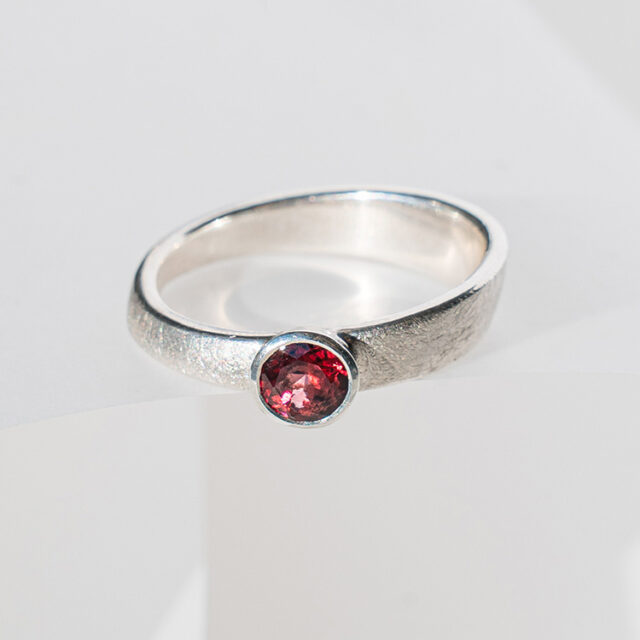 Silberner Ring mit dunkelrotem Stein auf hellem Hintergrund.
