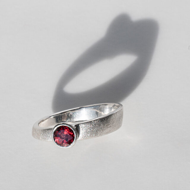 Silberner Ring mit dunkelrotem Stein auf hellem Hintergrund und mit Schattenwurf