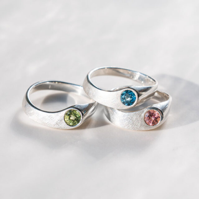 Drei Silberringe mit verschiedenen Edelsteinen in grün, blau und rosa liegen auf einem grauen Hintergrund.