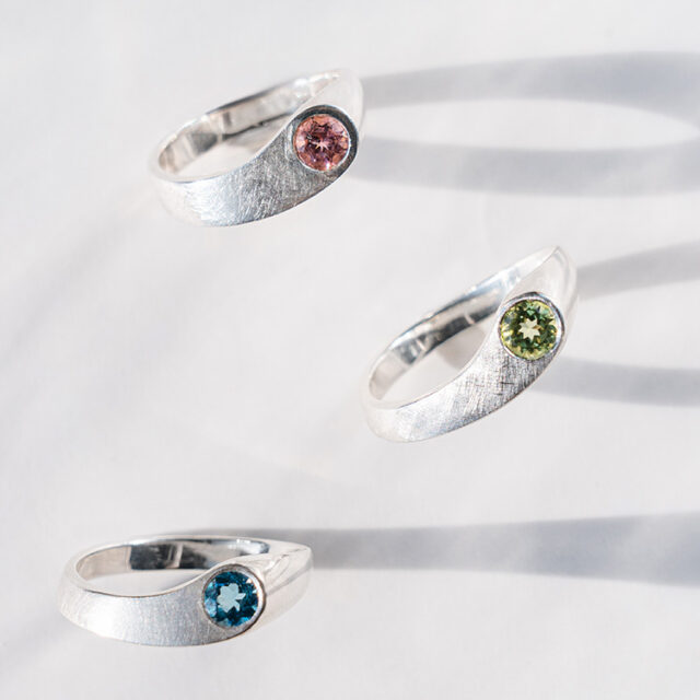Drei Silberringe mit verschiedenen Edelsteinen in grün, blau und rosa stehen auf einem grauen Hintergrund.