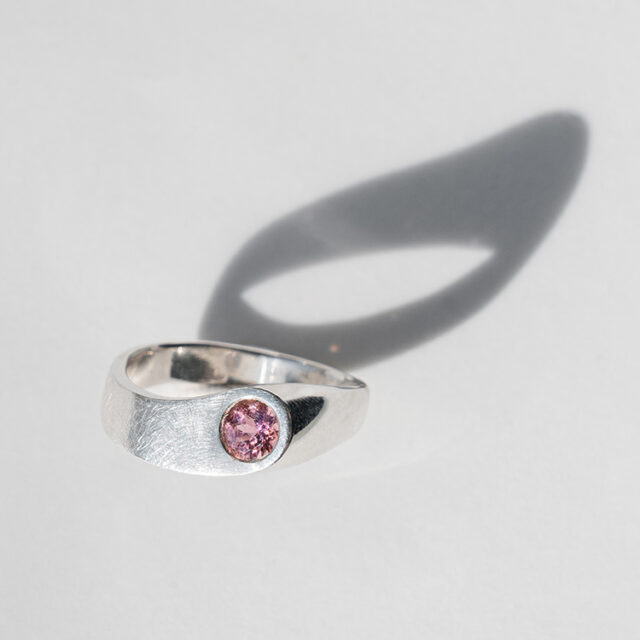 Silberring mit rosa Stein steht auf grauem Hintergrund.