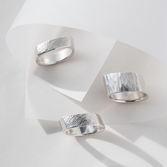 Drei Silberringe mit unterschiedlicher Oberflächenstruktur mit Kanten liegen auf einem grauen Hintergrund mit feinem Papier.