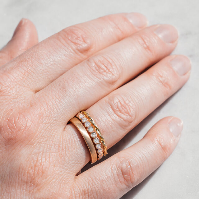 Hand mit drei Ringen am Ringfinger, ein gedrehter Kordelring, einer mit Brillanten und Millgriff Fassung und ein schlichter goldener Bandring.