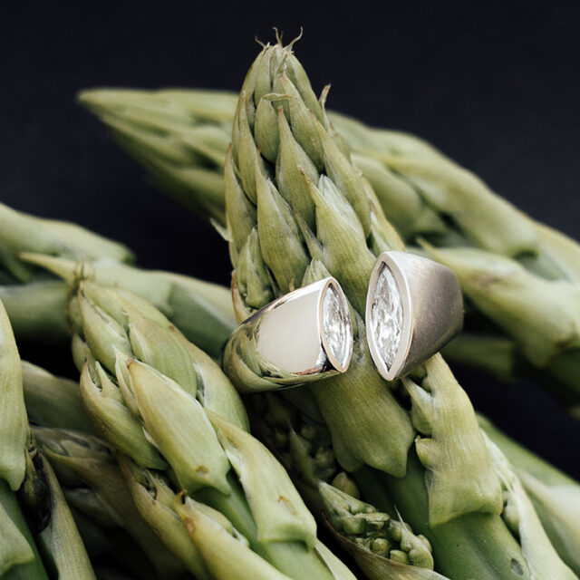 Ring aus Platin mit besonderer Form mit Spalt, in die Kanten wurde jeweils ein Diamant im Navette Schliff gefasst. Der Ring liegt auf Spargel.
