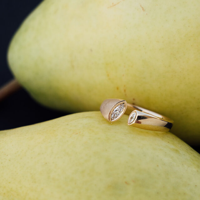 Goldener Ring mit besonderer Form mit Spalt, in die Kanten wurde jeweils ein Brillant im Navette Schliff gefasst. Der Ring liegt auf einer Birne.