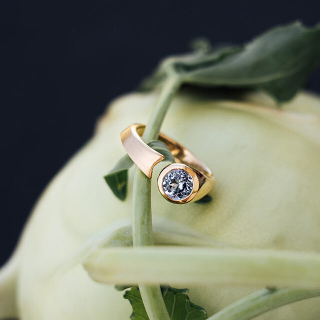 Goldener Ring mit Spalt und rundem Stein in hellem blau. Teilweise matt, teilweise glänzend. Der Ring liegt auf einem Kohlrabi.