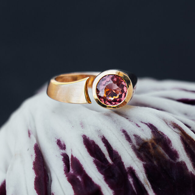 Goldener Ring mit Spalt und rundem Stein in dunklem rosa. Teilweise matt, teilweise glänzend. Der Ring liegt auf einem Radicchio.