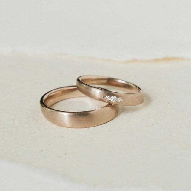 Ergonomische Eheringe aus Roséweißgold mit drei Brillanten im Damenring. Matte Oberfläche