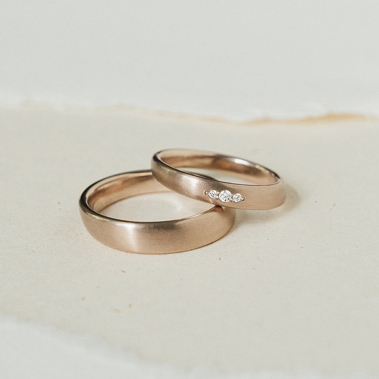 Ergonomische Eheringe aus Roséweißgold mit drei Brillanten im Damenring. Matte Oberfläche