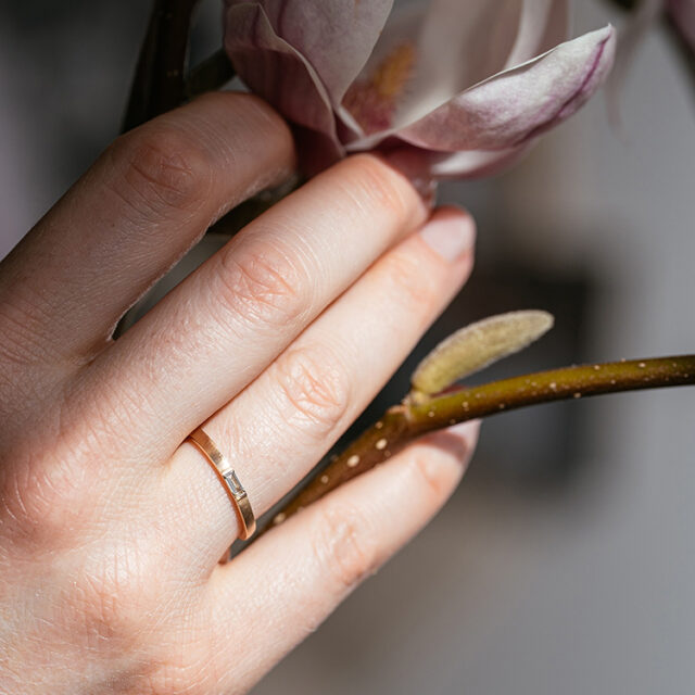 Ring in Roségold mit braunem Diamanten im Baguette-Schliff an einer Hand getragen, die eine Magnolie hält.