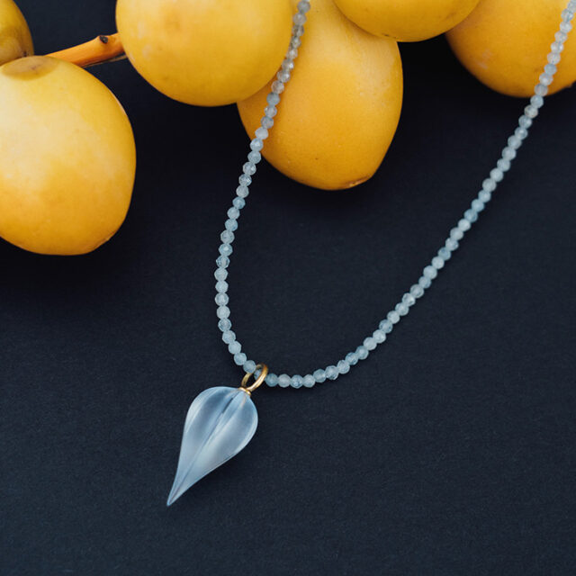 Anhänger aus 750er Gelbgold und einem synthetischen hellblauen Quarz an einer Aquamarinkette. Die Kette liegt auf schwarzem Untergrund neben frischen Datteln