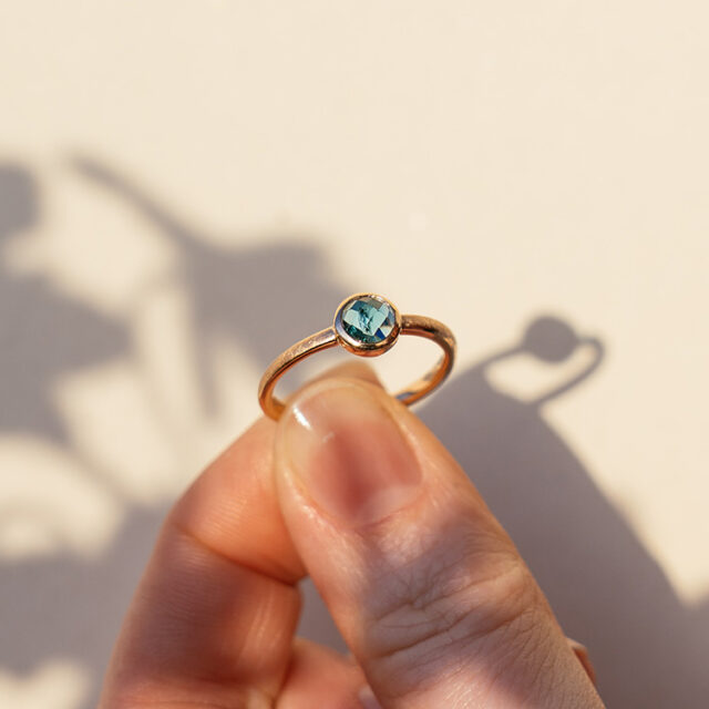 Ring aus Roségold mit rundem blauen Stein im Rosenschliff wird von zwei Fingern gehalten.