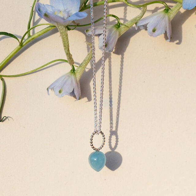 Eine silberne Kette mit Anhänger mit einem Ring aus Kugeldraht und Stein in Tropfenform in blau. Die Kette hängt in blauen Blumen