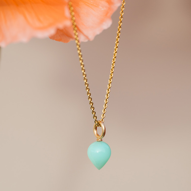 Goldene Kette mit Anhänger aus Gold und einem leuchtend Türkisen Stein in Tropfenform, hängend in einer Organgenen Blüte.