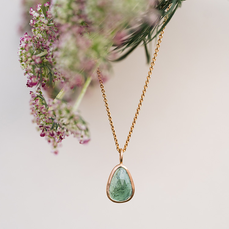 Kette aus Gold mit grünem Stein mit feinen Einschlüssen hängt in einer Blüte.
