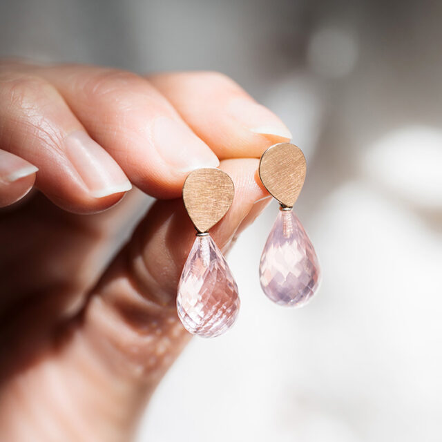Eine Hand hält zwei Ohrstecker mit einem Element aus Silber und Roségold mit Rosenquarzpampeln